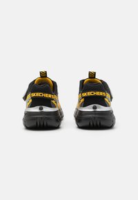 Zapatillas Skechers en negro y amarillo con parte superior de malla, acentos plateados y suela de goma. Cuentan con una asa en la parte trasera y una correa ajustable.