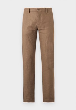 Homme portant un pull sans manches à rayures, un pantalon marron avec une ceinture noire et des sandales noires ouvertes au niveau des orteils, debout les mains dans les poches.