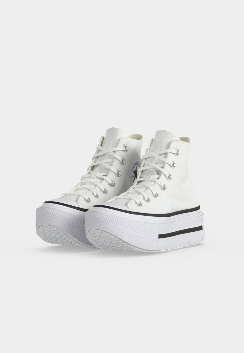 Paio di sneaker bianche alte con suola platform spessa, dettaglio a striscia nera e lacci bianchi su sfondo chiaro semplice.