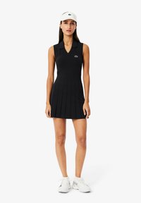 Abito polo senza maniche di colore nero con gonna plissettata, dotato di colletto e un piccolo logo verde sul petto. Un visiera bianca e scarpe da ginnastica completano il look.
