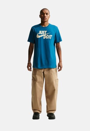 Uomo in piedi che indossa una maglietta Nike blu con il testo "Just Do It", pantaloni cargo beige, scarpe nere e tatuaggi visibili sulle braccia.