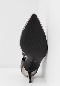 Chaussure à talon haut noire à bout pointu et talon aiguille fin, vue par-dessous sur fond blanc et angle de mur.