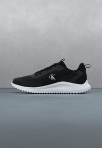 Baskets de sport noires avec semelle blanche et logo "cK" sur le côté, vues de profil sur un fond gris uni.