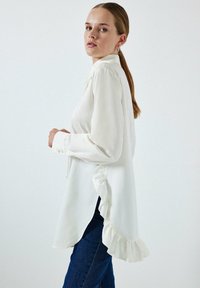 Camisa blanca de manga larga en tejido ligero con dobladillos y puños con volantes, que presenta un cuello clásico y un corte holgado, combinada con jeans azules.