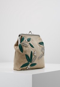 Beige stofclutch med metalramme, dekoreret med grønne broderede blade og sølvperleudsmykninger, stående på hvid overflade.