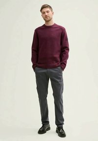 Young man standing with hands in pockets wearing a burgundy sweater, gray pants, and black shoes against a plain light background.