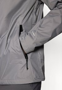 Pilkas vandeniui atsparus džemperis su užtrauktuku, elastinėmis rankovėmis ir elegantišku dizainu. Medžiaga turi tekstūruotą paviršių su reguliuojamu apačiu.