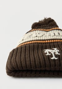 Barna kötött sapka bordázott mintával, fehér hímzett logóval és a tetején bojttal. Narancssárga és krémszínű csíkokkal díszítve.