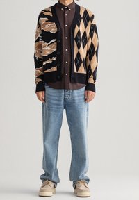 Stickad cardigan med svart, beige och tan-mönster; argyle- och vågmotiv. Lagd över en mörk skjorta, parad med blå jeans och sneakers.