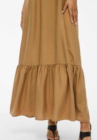 Brun maxiklänning med volangkant, tillverkad av textil med struktur. Har en avslappnad passform och en subtil lyster. Svarta sandaler kompletterar looken.
