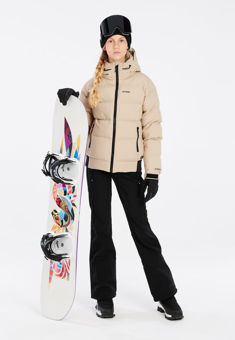 Beige Daunenjacke mit schwarzen Akzenten, schwarze Skihosen und Handschuhe. Hält ein buntes Snowboard mit aufwendigen Grafiken. Schwarze Skibrille.