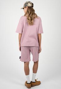 Femme portant un t-shirt rose oversize et un short assorti, une casquette camouflage, des chaussettes blanches et des chaussures bateau beige, debout de dos.