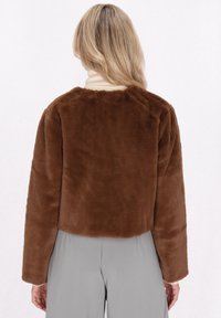Bruine faux-fur cropped jas met lange mouwen, ronde hals en een zachte textuur, gecombineerd met lichtgrijze broeken en een crèmekleurige coltrui.