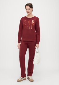 Burgundzka bluza z metalicznym logo "GAP", połączona z dopasowanymi spodniami. Obuwie w kolorze beżowym z czarnymi akcentami; nosi białą torebkę o textured.