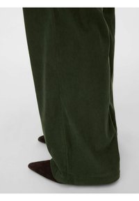 Zelené kordurové kalhoty s širokými nohavicemi, s texturovaným vzorem pruhů, dole vidět tmavé špičaté boty.