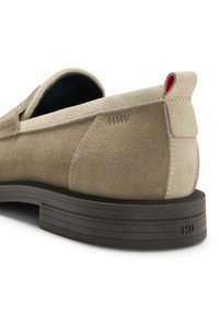 Beige Wildlederschlupfschuh mit gepolstertem Kragen, dicker schwarzer Sohle, roten Akzenten auf der Rückseite und dezenten Nähdetails im Obermaterial.
