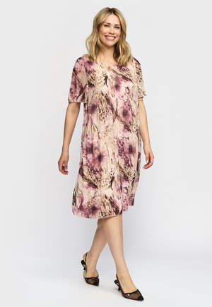 Kvinde smilende og gående iført en knælange, kortærmet blomsterkjole i pink og beige nuancer med sorte net-hæle.