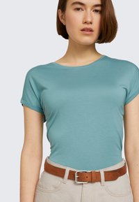T-shirt léger turquoise à manches courtes en tissu doux, avec un col rond. Associé à un pantalon beige et une ceinture en cuir marron.