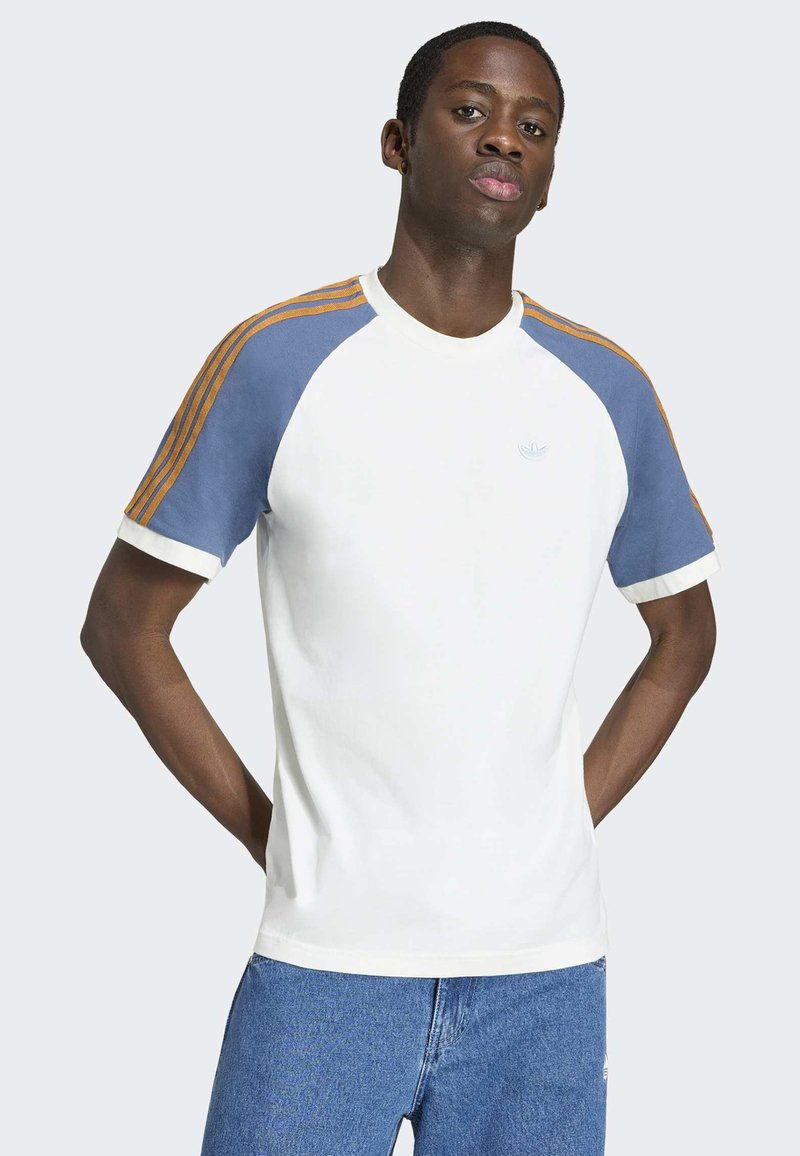 Junger Mann steht mit den Händen hinter dem Rücken, trägt ein weißes T-Shirt mit blau-orange gestreiften Ärmeln und blaue Jeans.