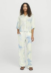 Dvoudílný outfit skládající se z košile s krátkým rukávem na knoflíky a kalhot s širokými nohavicemi. Světle žlutá látka s modrou batikou. Ploché sandály.