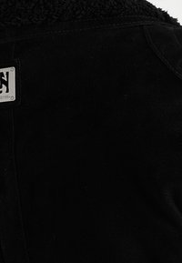 Giacca in suede nera con un colletto morbido e texturizzato. È visibile un'etichetta metallica che indica l'identità del marchio.