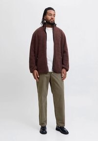 Brun fleecejacka med dragkedja och sidofickor, bärs över en vit t-shirt. Matchas med ljusolivgröna byxor och svarta läderskor.