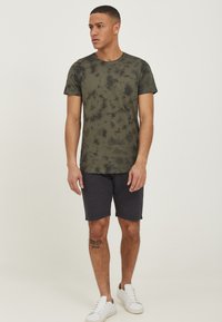 Majica s okruglim izrezom u maslinastoj boji s tie-dye dezenom, kratkih rukava, u kombinaciji s tamnim šortsem i bijelim tenisicama. Jednostavan i ležeran dizajn.