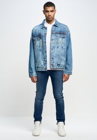 Chaqueta de denim azul claro con dos bolsillos frontales, cierres de botón y un ajuste relajado, llevada sobre una camisa blanca y combinada con jeans oscuros.