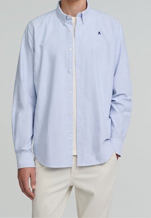 Homme portant une chemise boutonnée à fines rayures bleu clair et blanc sur un t-shirt blanc et un pantalon crème, main dans la poche.