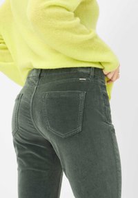 Grüne Cordhosen mit hoher Taille, zwei Gesäßtaschen und farblich abgestimmter Nähte. Kombiniert mit einem leuchtend gelben Pullover.