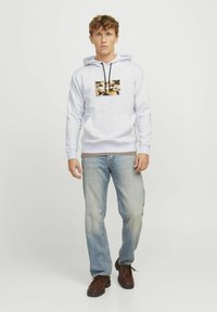 Sudadera gris con cordones negros y un parche rectangular de patrón. Llevada con jeans de mezclilla azul claro y zapatos marrones con cordones.