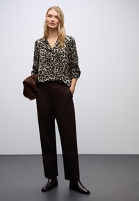 Blůza s leopardím potiskem, V-výstřih, dlouhé rukávy, spárovaná s hnědými kalhotami s vysokým pasem a tmavými kotníkovými botami. Hnědá kabelka přes rameno.