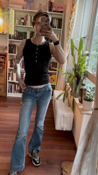 Svart ribbestrikket tanktopp med knappdetalj, kombinert med lyseblå flared jeans og mønstrede sneakers, satt i et rom med bokhyller.