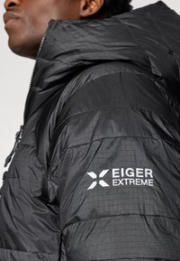 Casaco puffer preto com gola alta, fecho de zíper e bolsos laterais. Possui um acabamento texturizado com um logótipo bordado na manga.