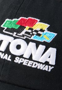 Μαύρο καπέλο με κεντημένες πολύχρωμες σημαίες αγώνων και λευκό κείμενο που λέει "Daytona International Speedway."