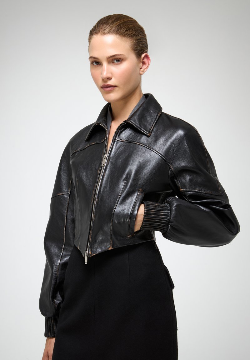 Chaqueta recortada de piel negra con un amplio cuello, cierre de cremallera y puños acanalados. Textura suave con detalles de costura en contraste.