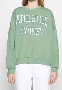 Felpa verde con spalle scese e orlo a costine. Presenta un testo bianco audace: "ATHLETICS SYDNEY 1987." Realizzata in morbido cotone.