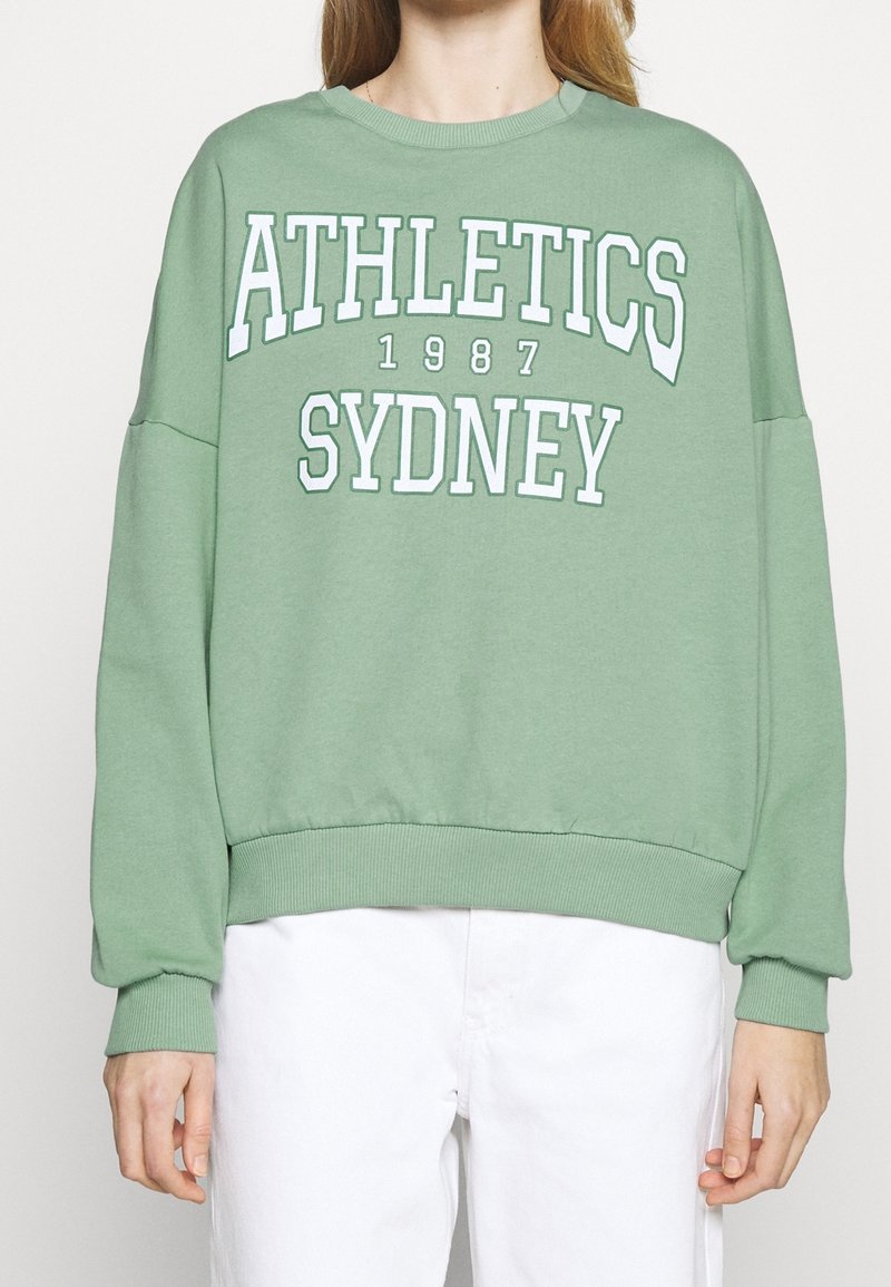 Felpa verde con spalle scese e orlo a costine. Presenta un testo bianco audace: "ATHLETICS SYDNEY 1987." Realizzata in morbido cotone.