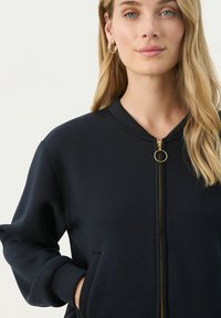 Kék zipzáros pulóver kerek nyakkal, elülső zsebekkel és arany árnyalatú cipzárral. Puha anyag sima felülettel.