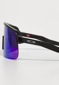 Gafas de sol negras con grandes lentes moradas degradadas y montura negra texturizada, que presentan un logo en forma de "O" en el lado y patillas inclinadas.
