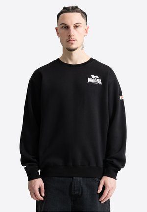 Mann mit geflochtenem Haar, trägt schwarzen Lonsdale London Sweatshirt mit Logo auf Brust und Ärmel, steht vor weißem Hintergrund.