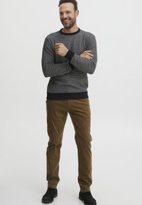 Suéter de punto gris jaspeado con puños de punto acanalado en navy, combinado con pantalones de pana marrones y botines negros, de pie en un fondo neutro.