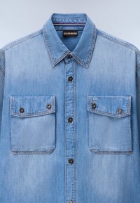 Hellblaues Jeanshemd mit durchgehendem Knopfverschluss, spitzem Kragen und zwei Brusttaschen mit Knopfklappen. Leicht ausgewaschene Textur.