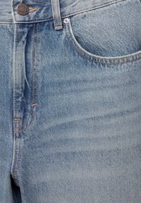 Jean en denim bleu clair en gros plan montrant la poche avant, un bouton en métal avec le nom de la marque embossé, une boucle de ceinture et des coutures détaillées.