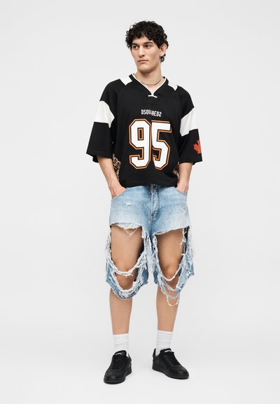 Νεαρός με σγουρά μαλλιά που φοράει μαύρη αθλητική φανέλα με τον αριθμό "95" και τζιν σορτς μέχρι το γόνατο με μεγάλες φθορές, στέκεται με τα χέρια στις τσέπες.