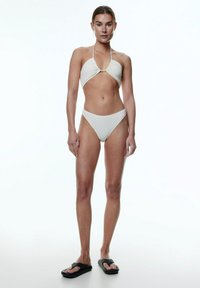 Weißes Schwimmbikini mit einem texturierten Haltertop in dreieckiger Form und hochgeschnittenem Höschen. Kombiniert mit schwarzen Flip-Flops.