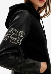 Czarna bluza z kapturem z ciałem z polaru i skórzanymi rękawami. "Mickey Mouse" w grubym białym napisie na lewym ramieniu. Posiada regulowany kaptur.
