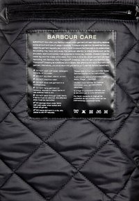 Zwart doorgestikt stoffen verzorgingslabel met witte tekst waarin wasinstructies, verzorgingsrichtlijnen en pictogrammen voor stofonderhoud worden beschreven.