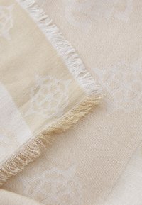 Vaaleanbeige huivi, jossa on pehmeä rakenne, hienovarainen kuvio ja hapsureunat. Materiaali heijastaa hieman valoa, mikä parantaa sen ulkonäköä.