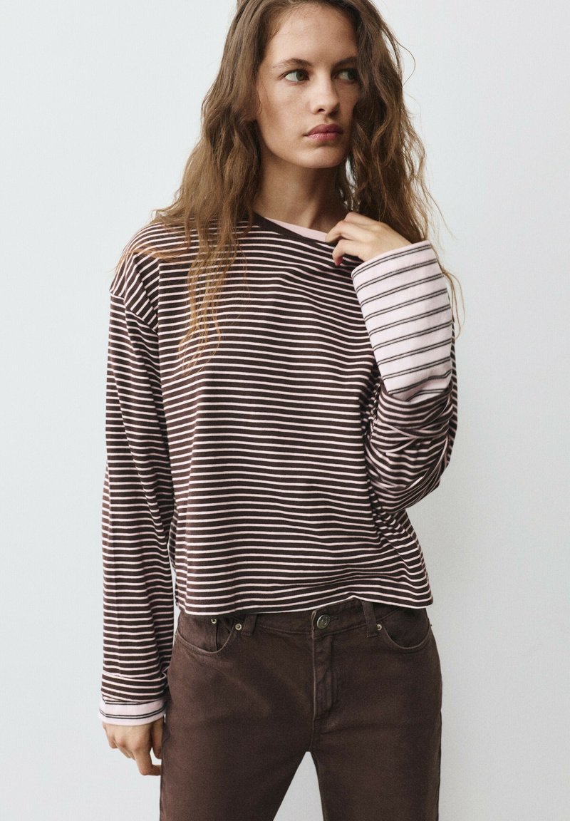 Femme aux longs cheveux ondulés portant une chemise à manches longues rayée marron et blanche et un pantalon marron foncé, regardant sur le côté avec une main près du cou.
